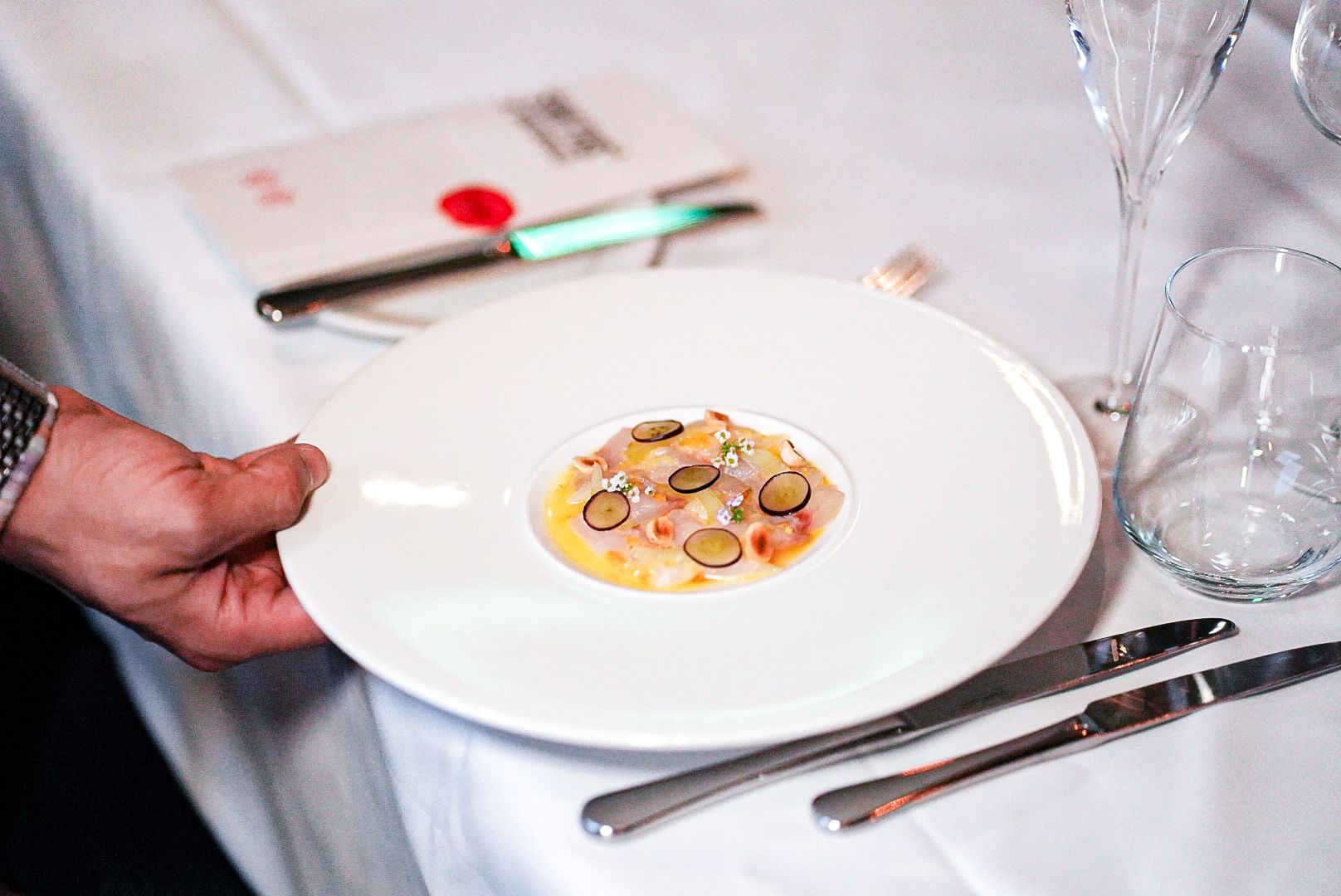 A waiter provides professional service by presenting the cured sea bream dish to guests in the private dining room during a 7-course tasting menu meal.